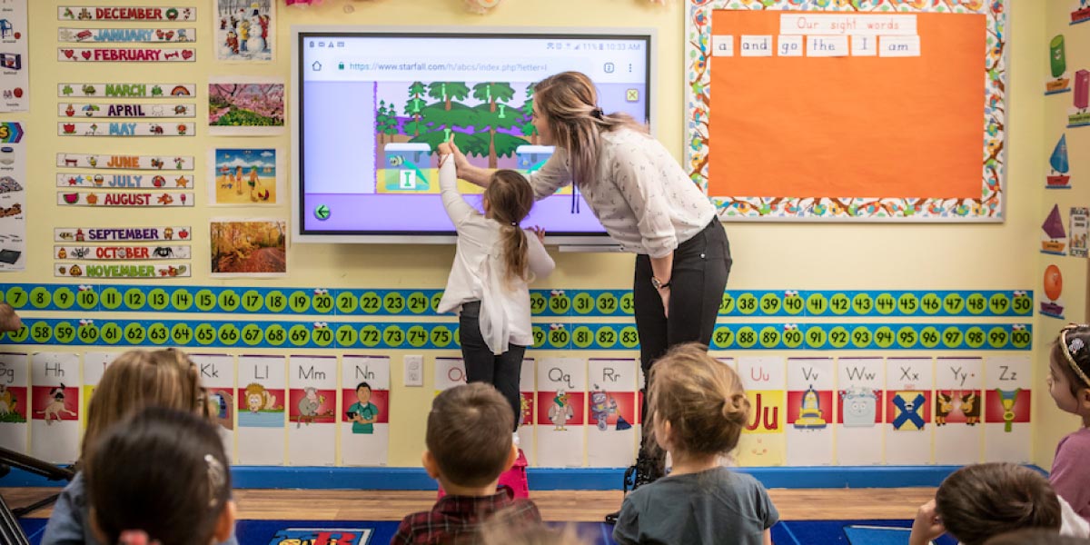In una classe di bambini un’insegnante guida un’alunna nello svolgimento di attività sulla lavagna multimediale interattiva