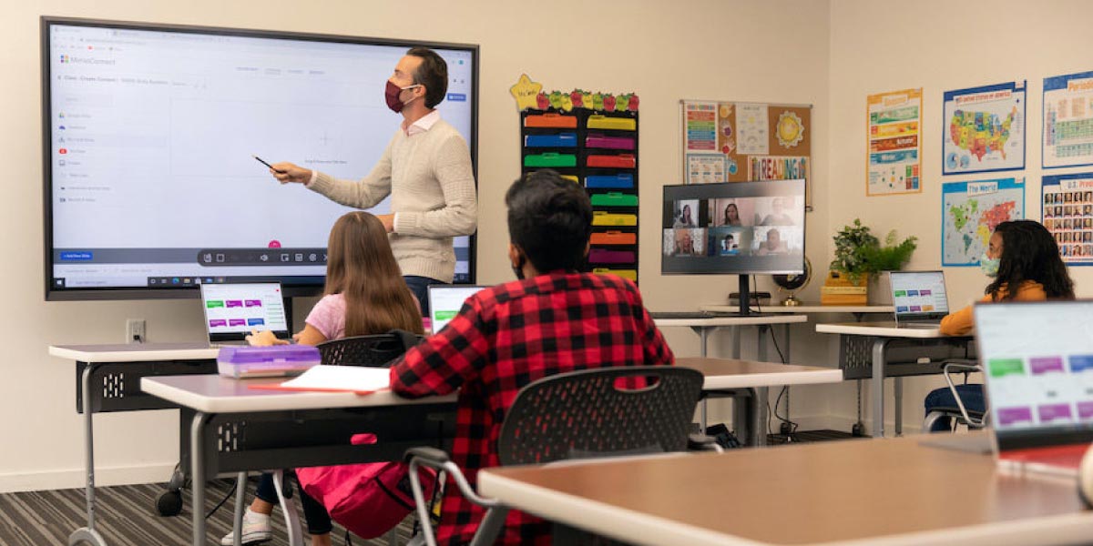 Un professore insegna agli studenti in classe mentre indica una lavagna multimediale interattiva e su un display si vedono altri alunni connessi da remoto
